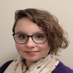 Aleeh Schwoerer headshot. Aleeh has short light brown curly hair. She is wearing black glasses and a white floral scarf.